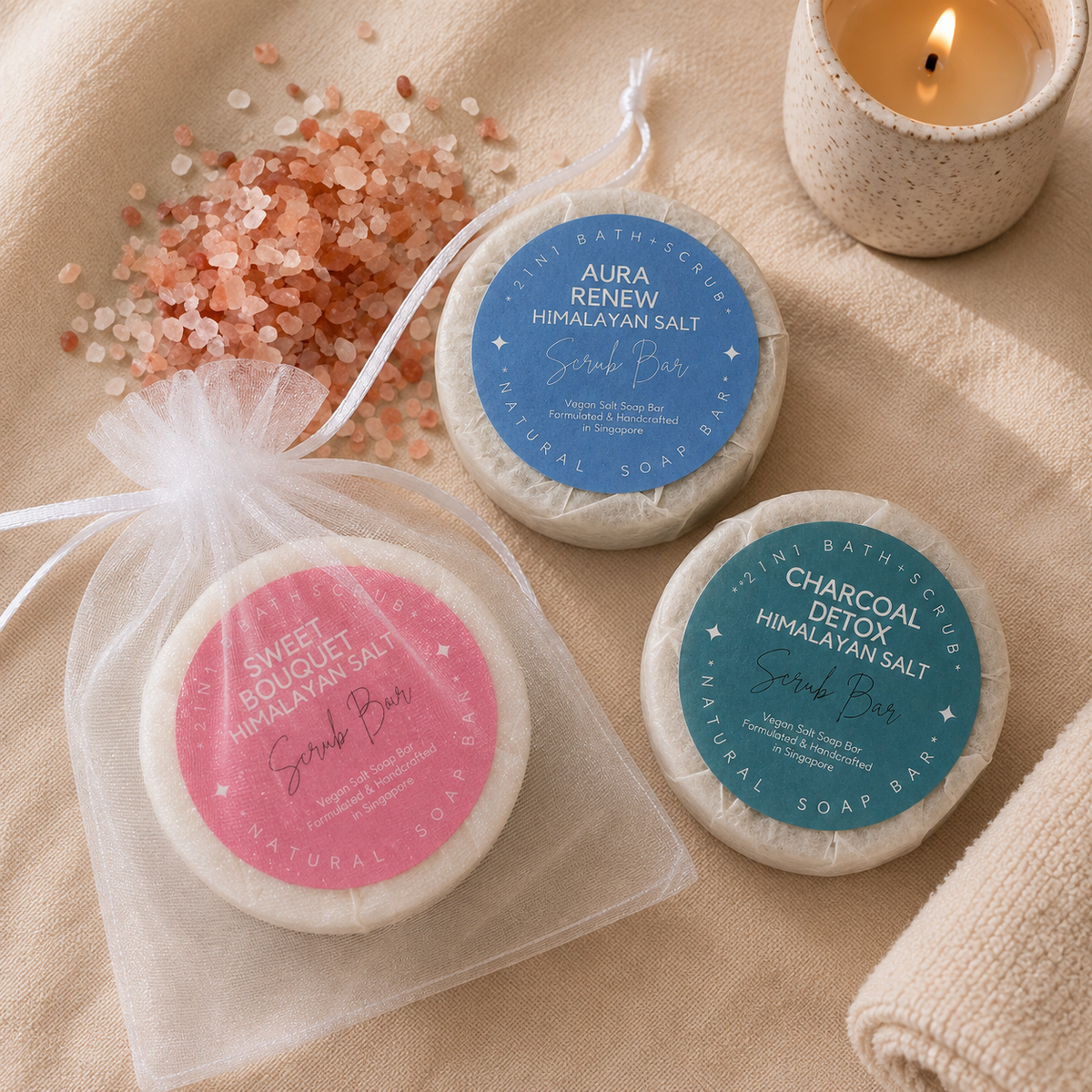 Three round natural soap bars with colorful circular labels are arranged on a beige fabric surface beside a white sheer drawstring bag, pink salt crystals, and a lit candle. The labels read Sweet Bouquet Himalayan Salt, Aura Renew Himalayan Salt, and Charcoal Detox Himalayan Salt, with bath series text visible on the top of each bar.