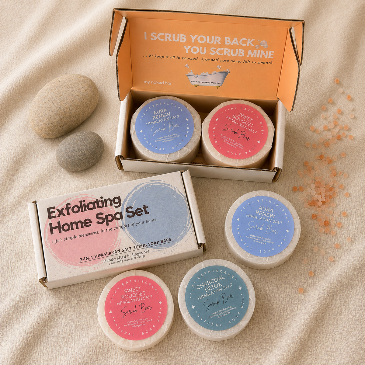 My Naked Bar exfoliating home spa set containing two 2-in-1 scrub bars in an open cardboard box, with a matching closed boxes and three loose salt scrub soap bars arranged on a beige fabric background with decorative stones and salt crystals.