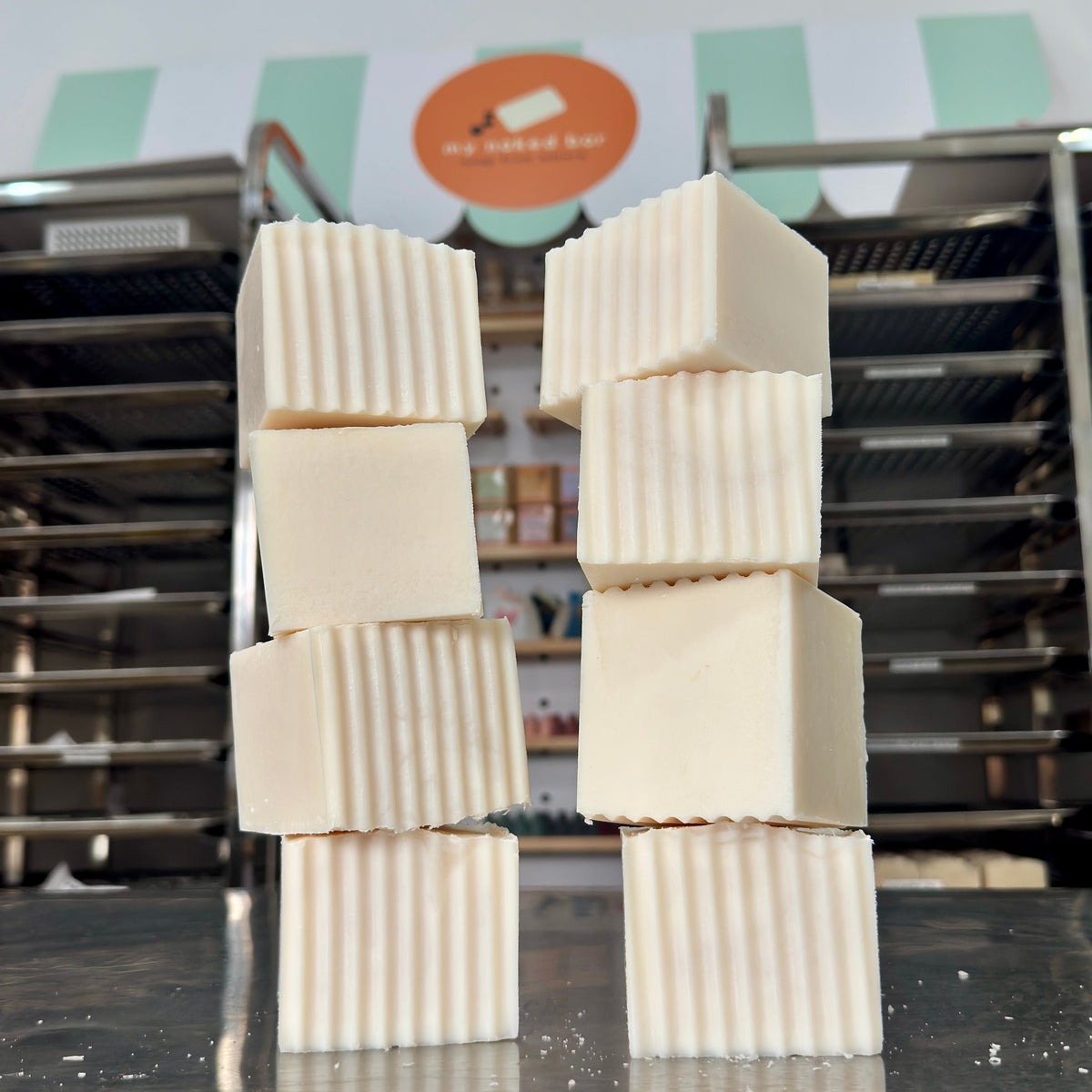 Four stacked My Naked Bar rectangular bars of cream-colored handmade soap for kitchen dishes with vertical ridges, standing on a stainless steel table with a blurred soap workshop background.