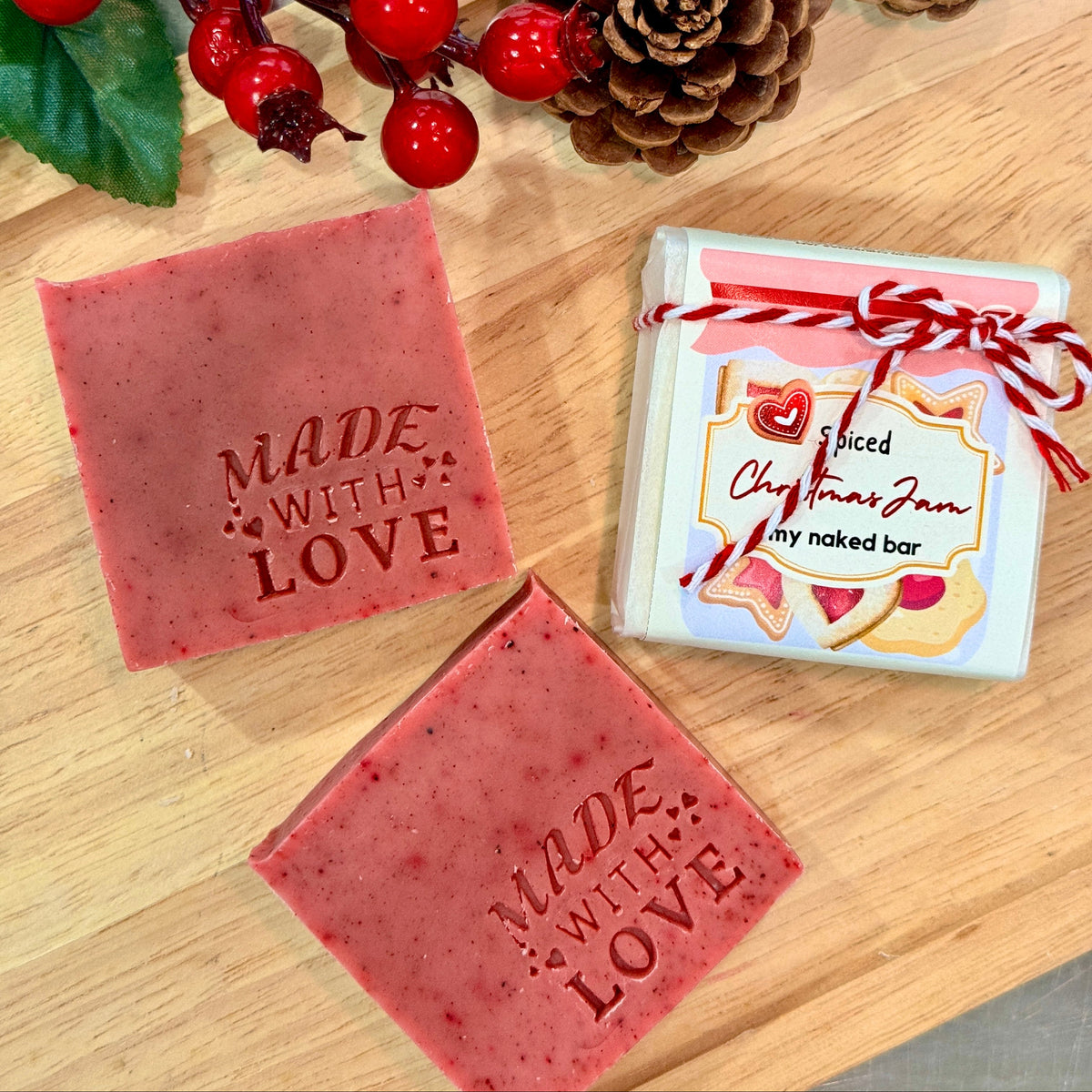 Three handmade pink soap bars are arranged on a wooden board with decorative red berries and pinecones. One soap bar is embossed with the words 'MADE WITH LOVE,' and the packaged soap in the center reads 'Spiced Christmas Jam my naked bar.