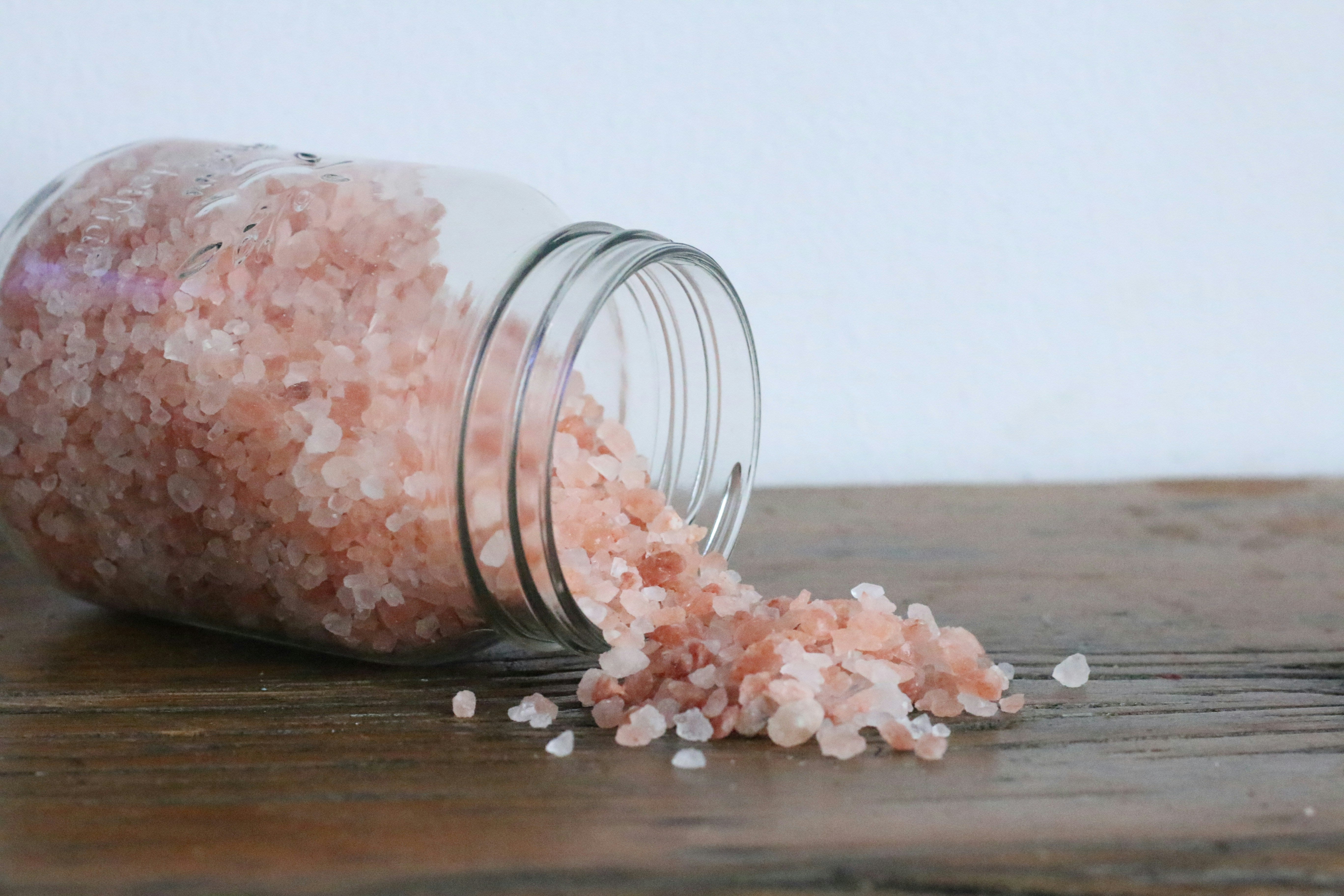Glass jar lying on the side with coarse Himalayan salt pouring out onto a wooden table