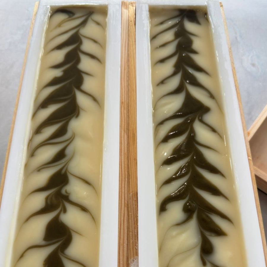 Two rectangular logs of soap bars in white molds with a marbled swirl pattern of cream, beige, and green, set in a wooden tray.