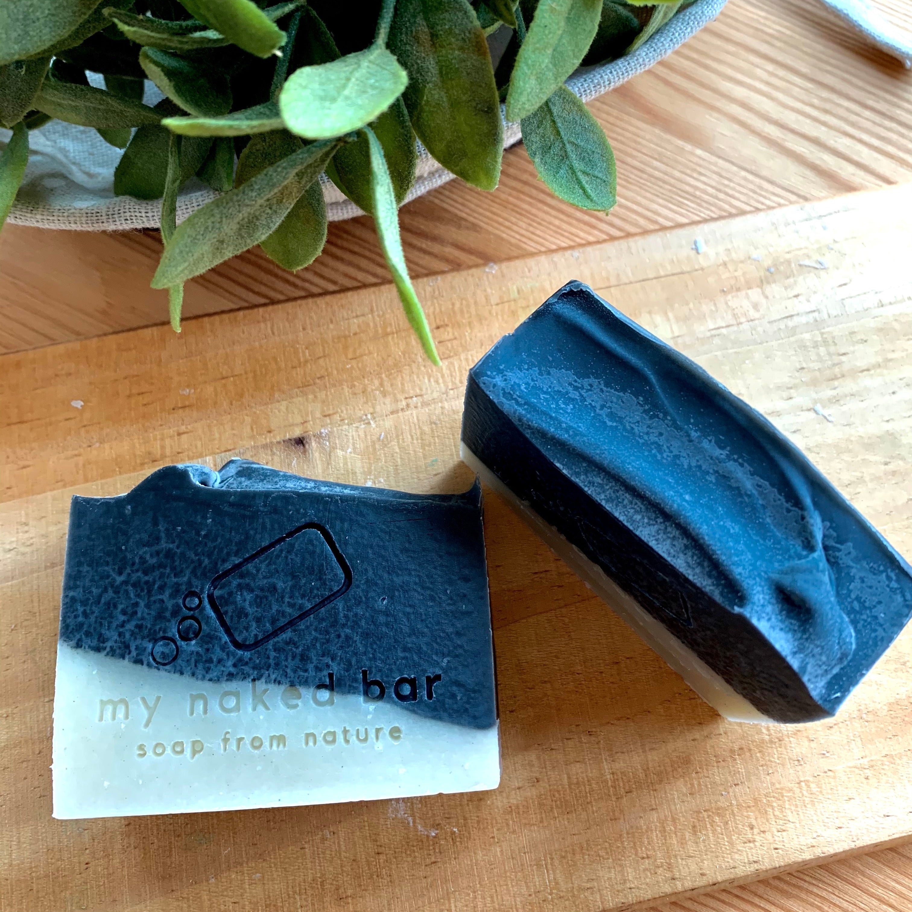 2 bars of Black Mojo soap bar one front facing and other top facing on a wooden board with plant in the background