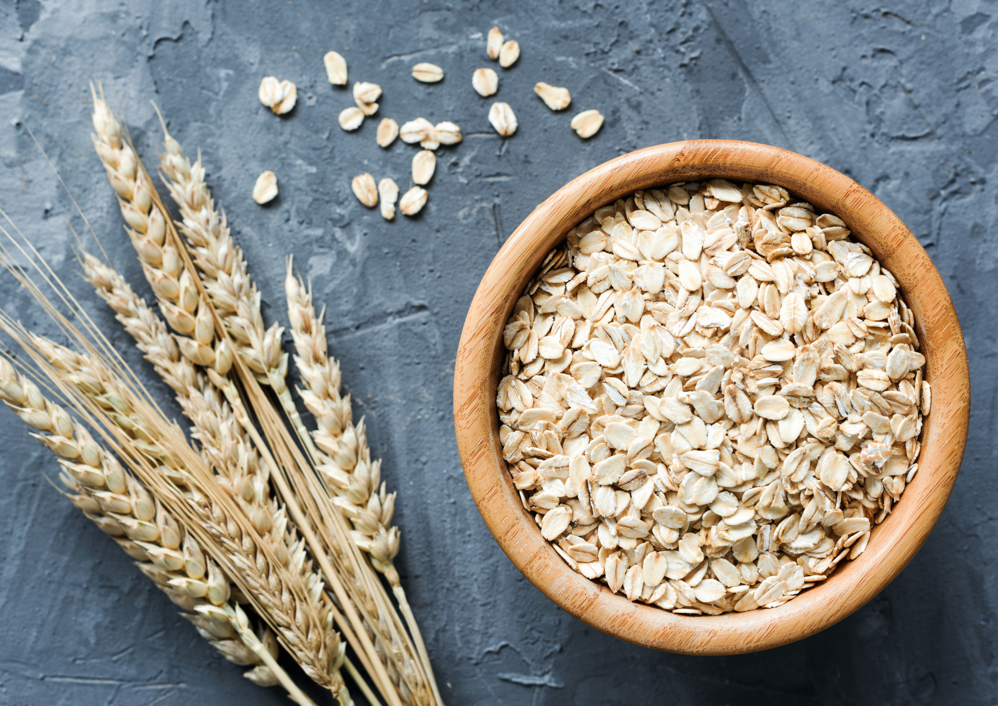 Top view of oats in a wooden bowl with the plant beside against a rugged grey slate