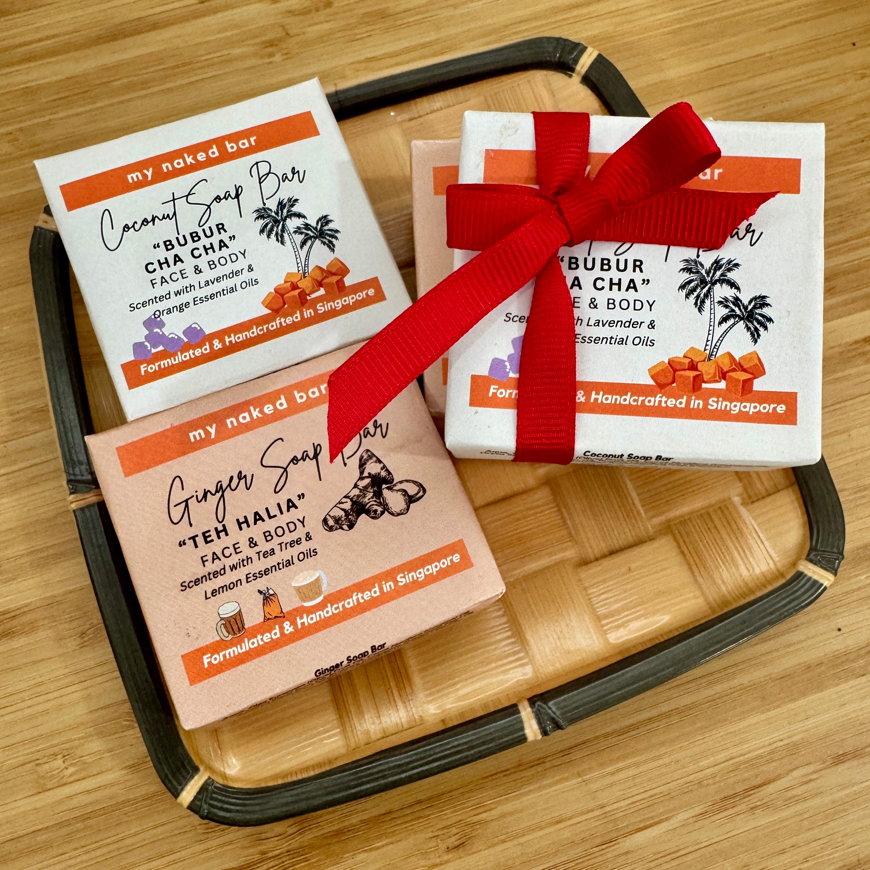 Bubur Cha Cha Coconut Soap Bar and Teh Halia Ginger Soap Bar lying face up on a bamboo late, beside the pair bundled up together in a red ribbon as Singapore Soap Gift Bundle