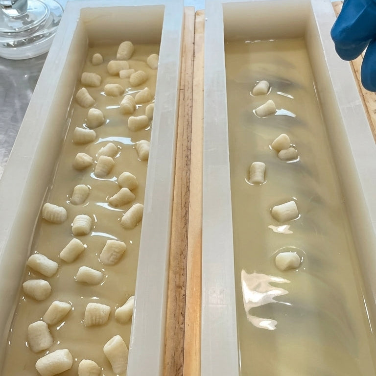 Two rectangular soap mould filled with soap batter with a gloved hand sprinkling soap dough shaped in marshmallow on the top.