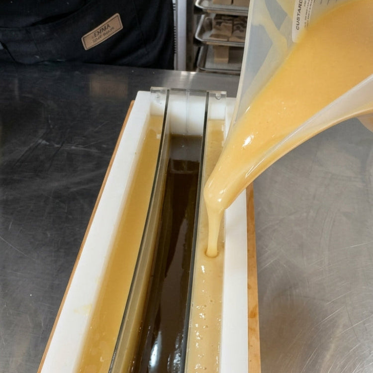 Crafting of Pine Fir Yule soap bar where the soap batter is being poured from a measuring cup into a long rectangular mold on a stainless steel workbench.